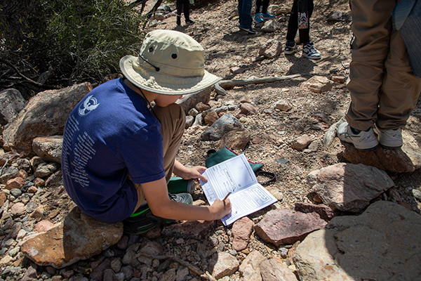 A boy crouches down on the trail writing in a journal