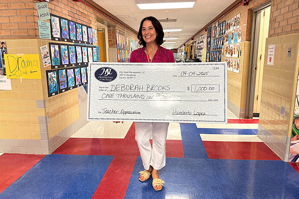 A woman in a pink shirt and white pants smiles as she holds a giant check in a school hallway