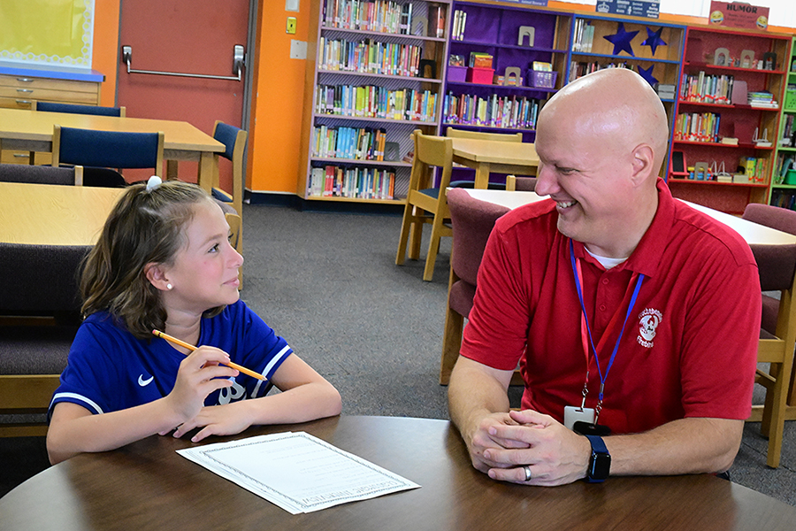 A girl in a blue shirt sits at a table in the library with a man in a red shirt
