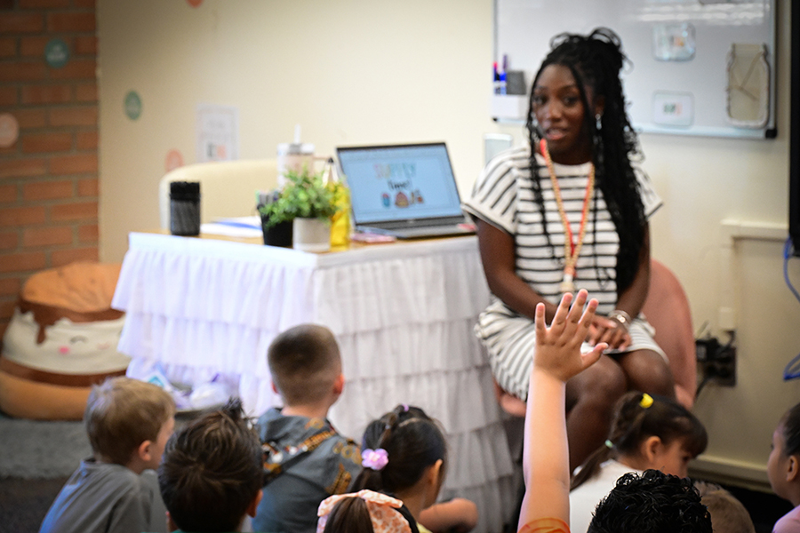 A teacher in a white striped dress addresses her class as a student raises their hand in the foreground